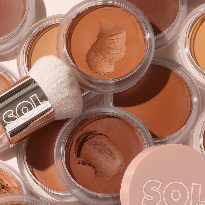 ColourPop SOL Face & Body Bronzing Balm in Medium sits open with a creamy swipe, surrounded by additional warm-toned shades and a SOL-branded makeup brush on a light background, highlighting the warm neutral tone.