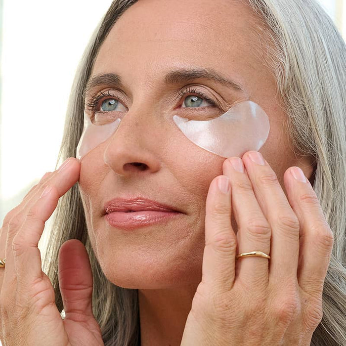 Mature woman with grey hair applying white Patchology eye gels for a brightened, refreshed undereye area.
