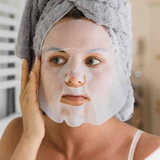Woman wearing a white sheet mask and grey towel wrap during a skincare routine.