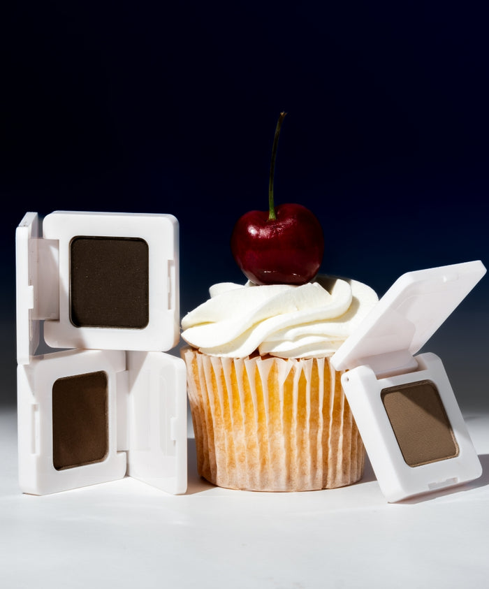 Three open compacts of Back2Brow Powder in natural brown shades arranged around a cupcake topped with white frosting and a red cherry against a dark background.
