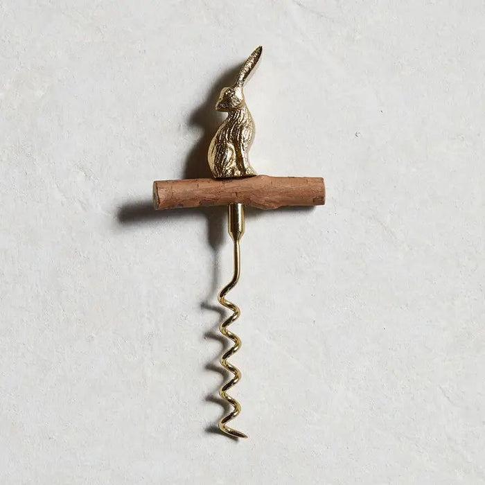 A bottle of wine with a golden foil sits beside the BE HOME - HARE CORKSCREW by BE HOME, INC, featuring a brass rabbit-shaped handle and wooden grip on a light wood surface—an elegant gift for any hostess.