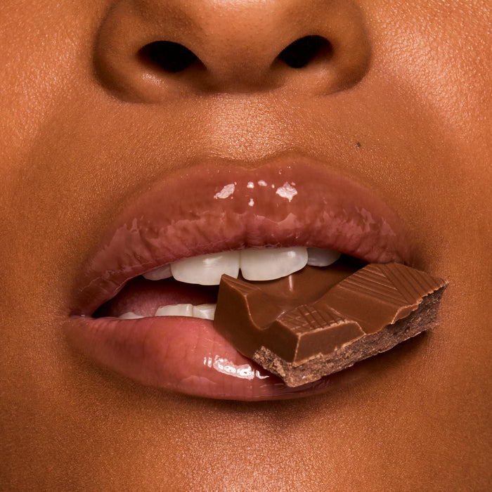 Glossy, cocoa-tinted lips wearing ColourPop Fresh Kiss Glossy Balm in Chocolate Milkshake hold a square of milk chocolate between teeth in a macro close-up, showcasing high-shine, cushy hydration and a creamy nude tint.