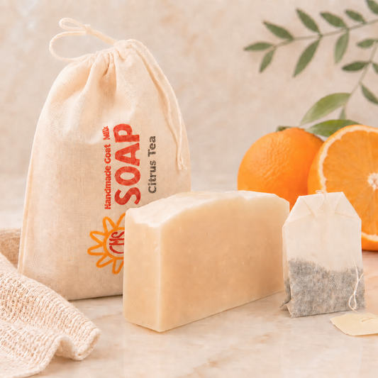 Bar of goat milk soap, with tea bags, and a drawstring bag on a light surface with oranges and leaves in the background.