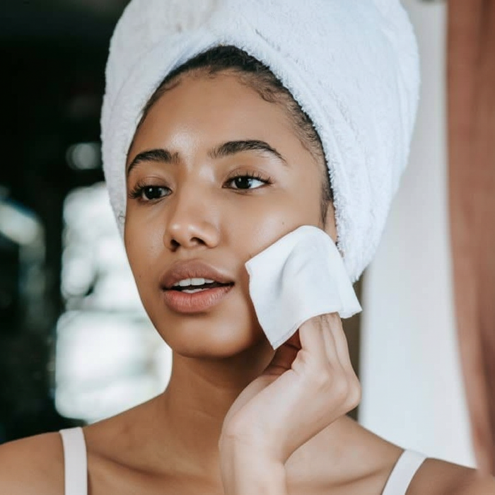 Woman with a white towel wrap gently cleansing her cheek with a soft cotton pad.