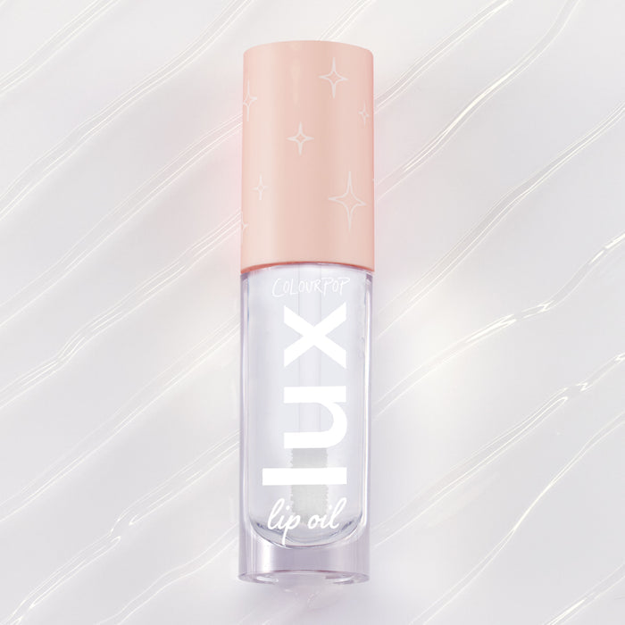 ColourPop Dew Drop Lux Lip Oil lying flat, a clear high-shine formula in a rectangular tube with blush-pink cap printed with starbursts and bold lux lettering, set on a pearlescent swirled backdrop for a glossy finish.