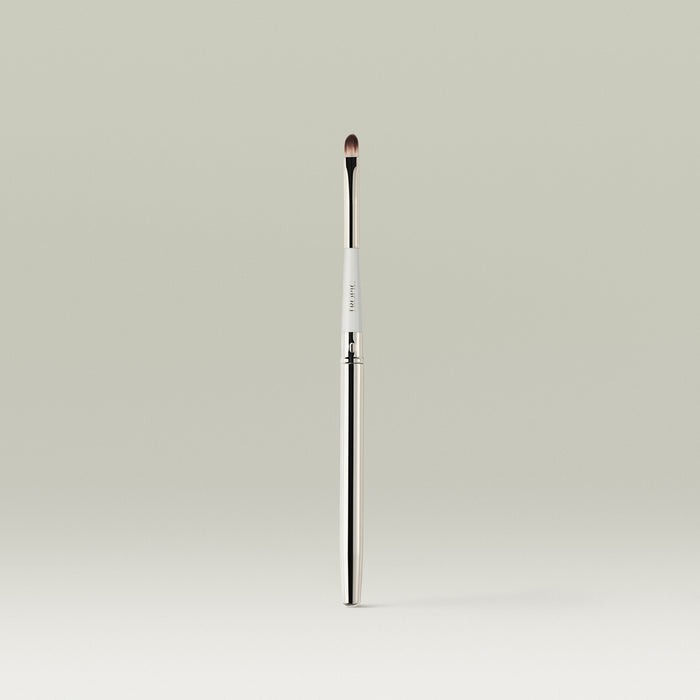 A sleek, dual-ended lip brush with a slim metallic handle and a small, rounded bristle tip, standing upright against a soft neutral background.