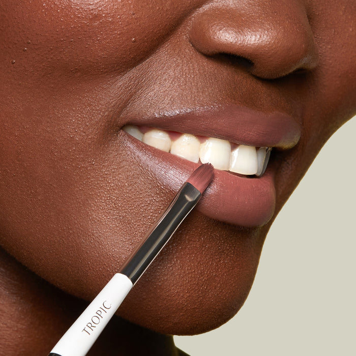 A close-up of a person’s lips as a lip brush applies a smooth, mauve lip colour. The brush, labelled “Tropic,” is gently defining the edge of the lower lip.