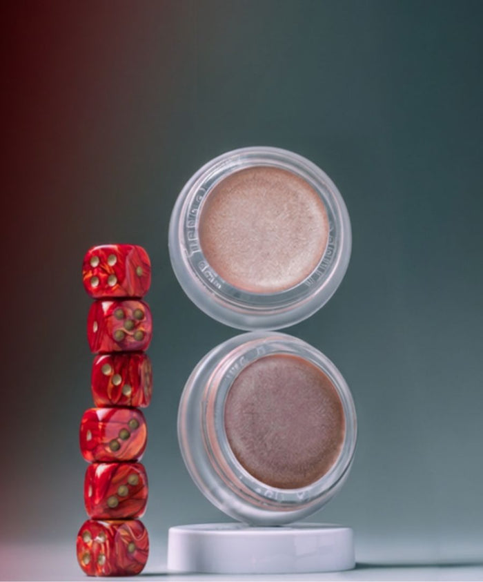 Two open jars of Luminizer cream makeup with a stack of red dice beside them on a smooth surface against a muted dark background