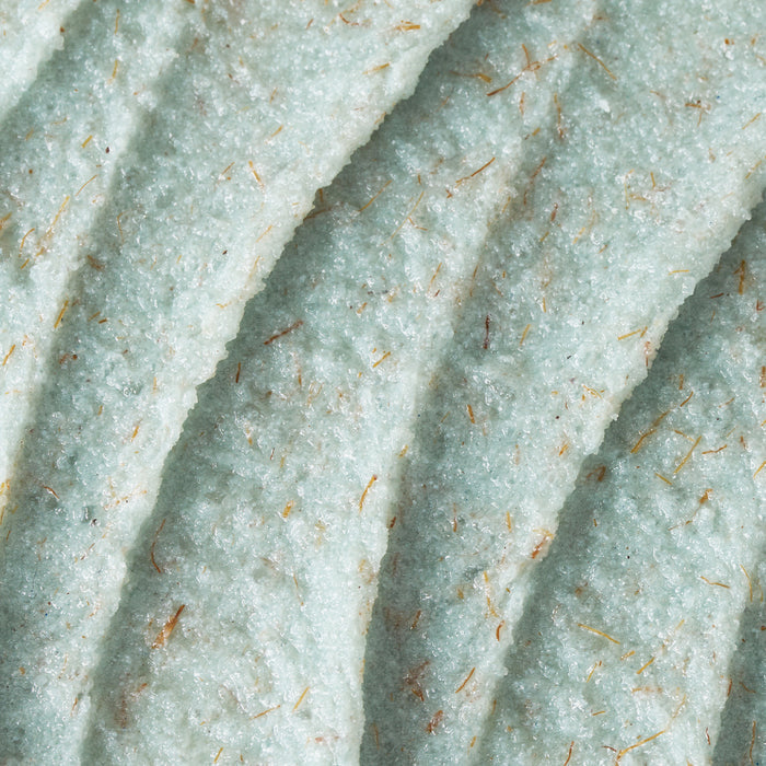 A close-up texture shot of Foot Smooth Enzymatic Scrub, showing its pale mint-green formula spread in soft, sculpted ridges. The scrub has a creamy yet grainy consistency, with visible natural exfoliating particles dispersed throughout, creating a gently buffing, skin-polishing texture.