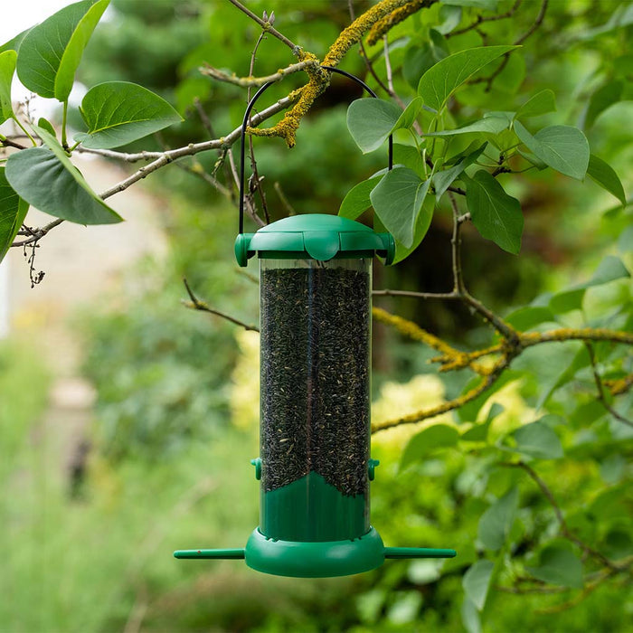 A green Gardman flip top nyjer seed feeder hanging from a branch, filled with nyjer seeds, surrounded by green foliage.