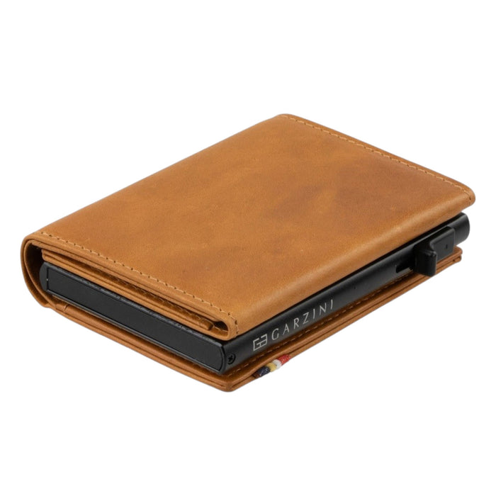 The GARZINI Slim Trifold RFID-blocking leather wallet in cognac features a metal card holder and coin pocket, with a black card partially visible on the side, photographed on a white background.