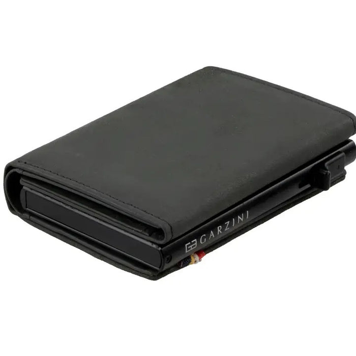 A GARZINI Slim Trifold RFID-blocking leather wallet in carbon black is shown closed, featuring a minimalist design, metal card holder, coin pocket, and the "GARZINI" logo on the side.
