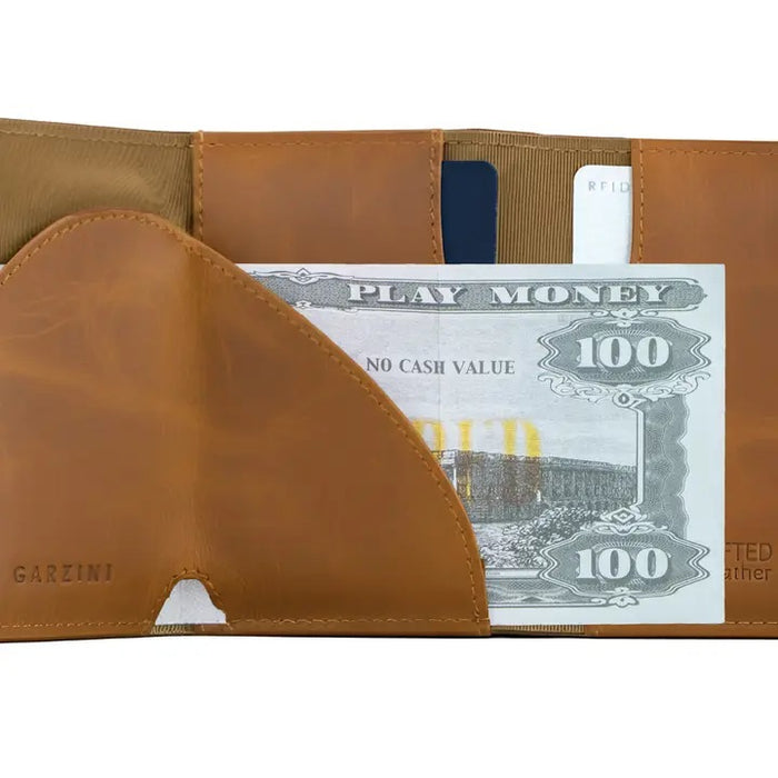 The GARZINI Slim Trifold RFID-blocking leather wallet in cognac features a metal card holder and coin pocket, with a black card partially visible on the side, photographed on a white background.