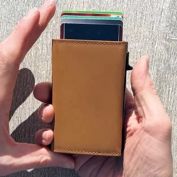 The GARZINI Slim Trifold RFID-blocking leather wallet in cognac features a metal card holder and coin pocket, with a black card partially visible on the side, photographed on a white background.