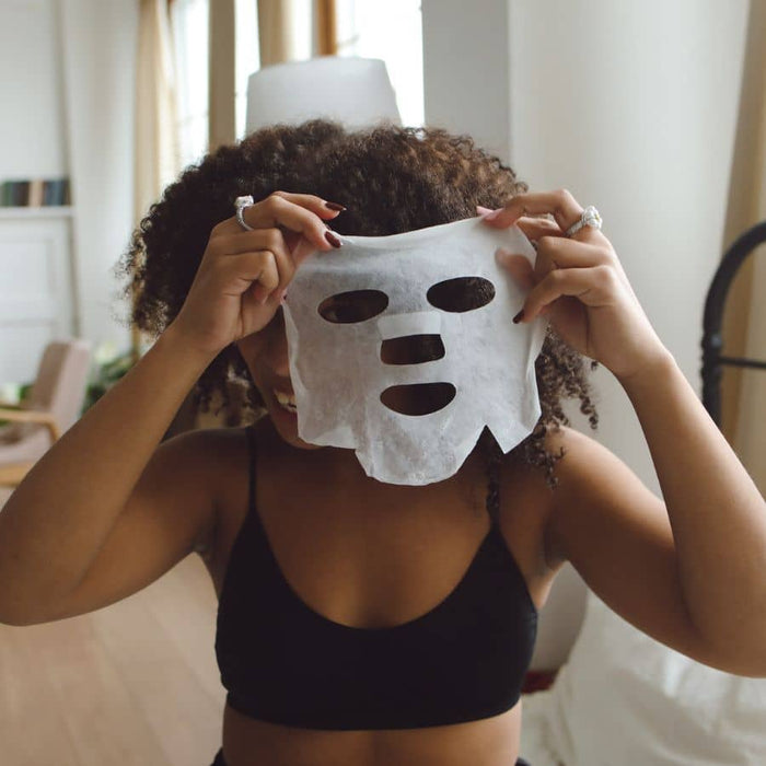 Woman with curly hair holding up a white fiber sheet mask in front of her face during a home skincare session.