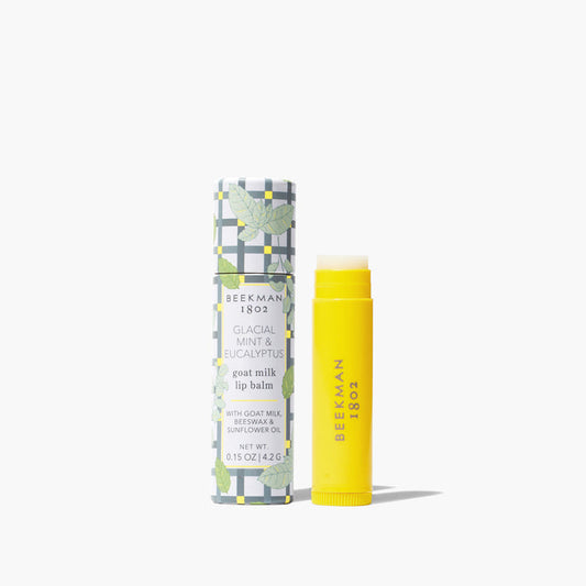 Yellow uncapped tube of Beekman 1802's Glacial Mint & Eucalyptus lip balm standing next to the packaging tube for the lip balm that is designed with leaves and gingham pattern, on a white background.
