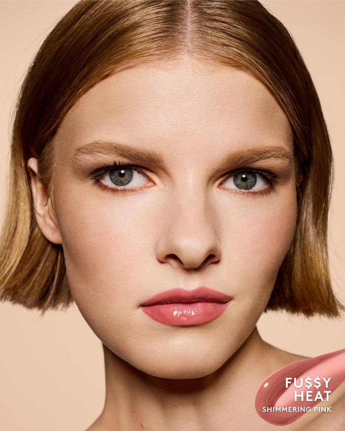 Close-up of a woman with short auburn-blonde hair wearing a glossy shimmering pink lip product. The bottom-right corner includes a pink swatch labeled 'FUSSY HEAT SHIMMERING PINK'.