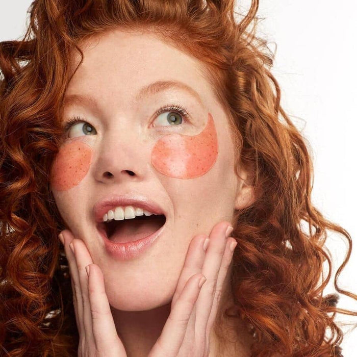 Surprised redhead woman with curly hair wearing orange, speckled hydrogel eye patches for a bright and energized skincare routine.
