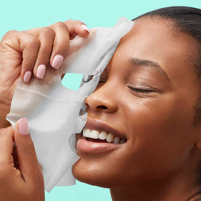 Smiling woman applying a white sheet mask against a mint background.