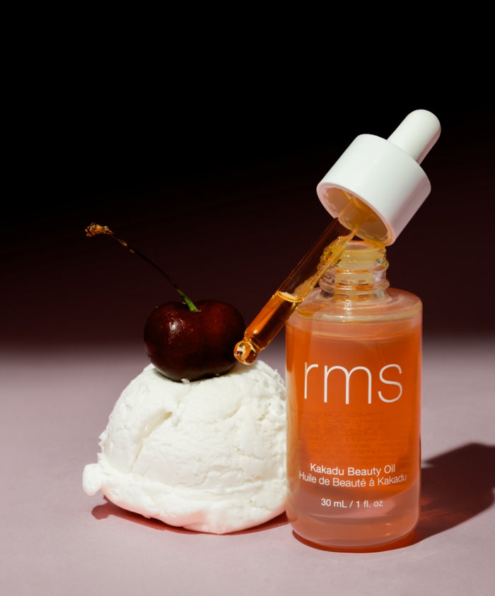 Bottle of Kakadu Beauty Oil with dropper releasing oil, placed next to a cherry resting on a scoop of white cream, highlighting clean beauty and natural ingredients.