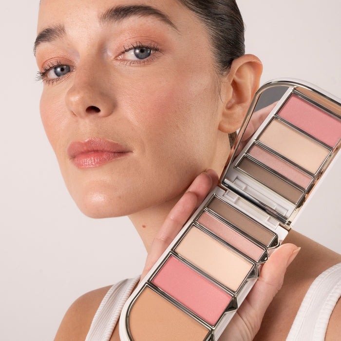 A person with natural makeup holds the Tropic Colour Palette in the “Spring” shade near their face, showing the soft pink, nude, and brown tones applied to their skin.