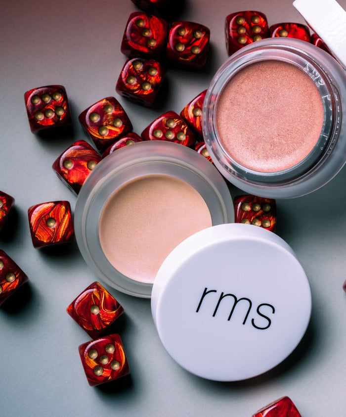 Open containers of Luminizer cream makeup in soft pink and beige shades surrounded by decorative red dice with gold dots against a gray background