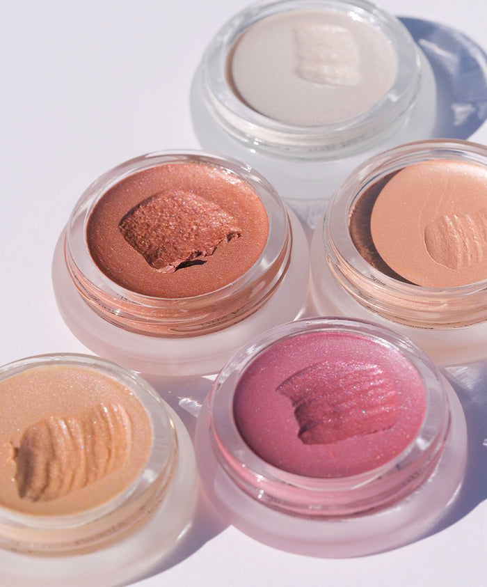 Five open jars of creamy, shimmering makeup in warm and neutral tones, showcasing the texture of Luminizer in natural light on a white surface.