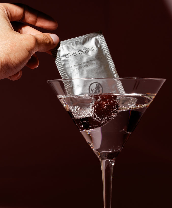 Hand holding a silver foil packet labeled rms beauty being dipped into a martini glass filled with clear sparkling liquid and a dark cherry, showcasing Makeup Remover | Ultimate Makeup Remover Wipe in a clean beauty setting.
