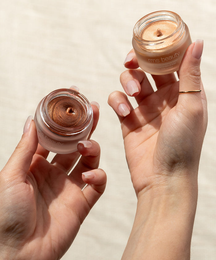 Two hands holding open jars of glowing cream makeup showcasing natural, radiant shades of RMS Beauty's Master Radiance Base with clean, sustainable packaging.