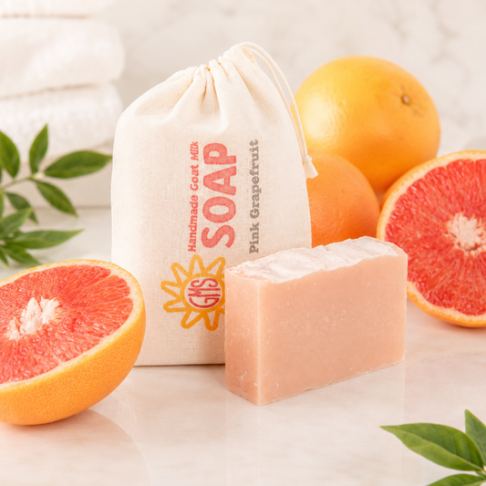 Bar of goat milk soap with a pink grapefruit theme, surrounded by pink and orange grapefruits and a drawstring bag labeled 'Pink Grapefruit Soap'.