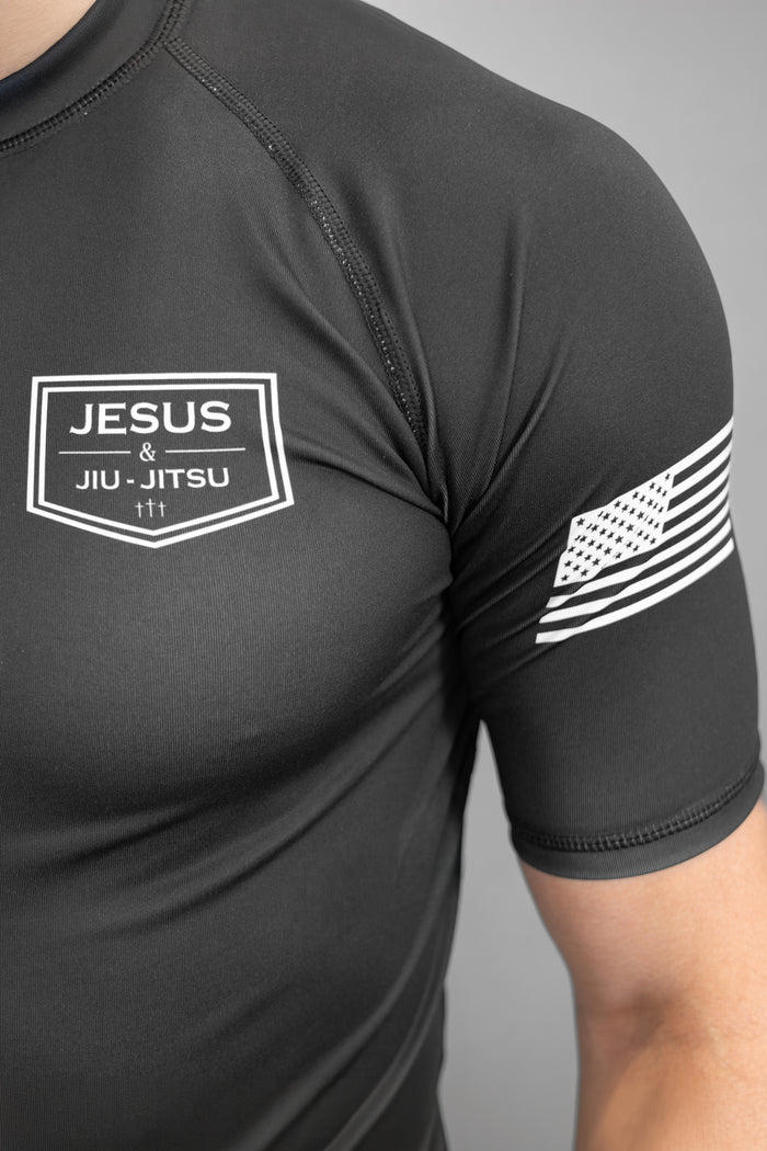 Black athletic shirt with 'Jesus & Jiu-Jitsu' logo and American flag sleeve design.