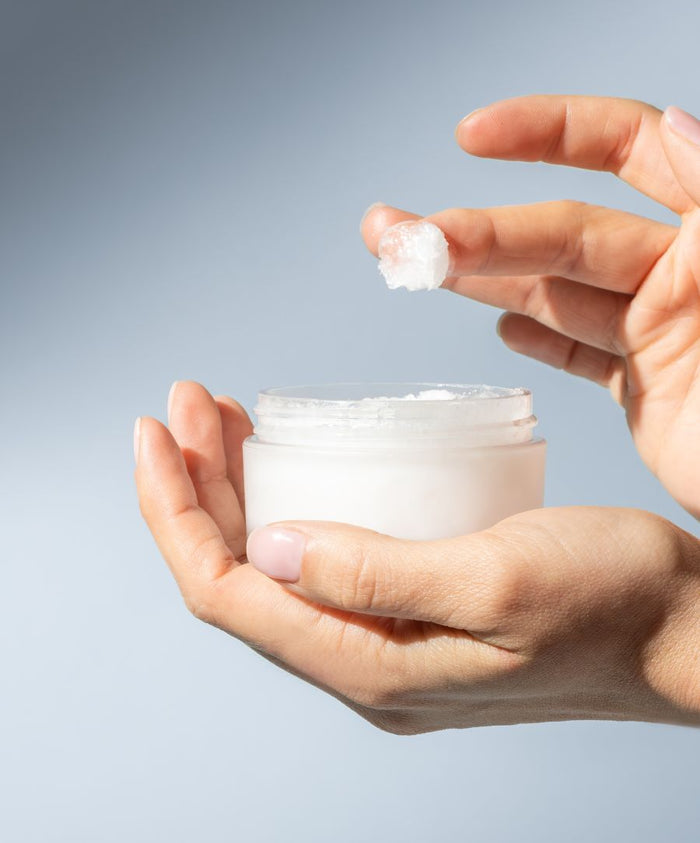 Hands holding a jar of Raw Coconut Cream with a small amount scooped onto fingers, showcasing its creamy, natural texture for skincare and body care uses.