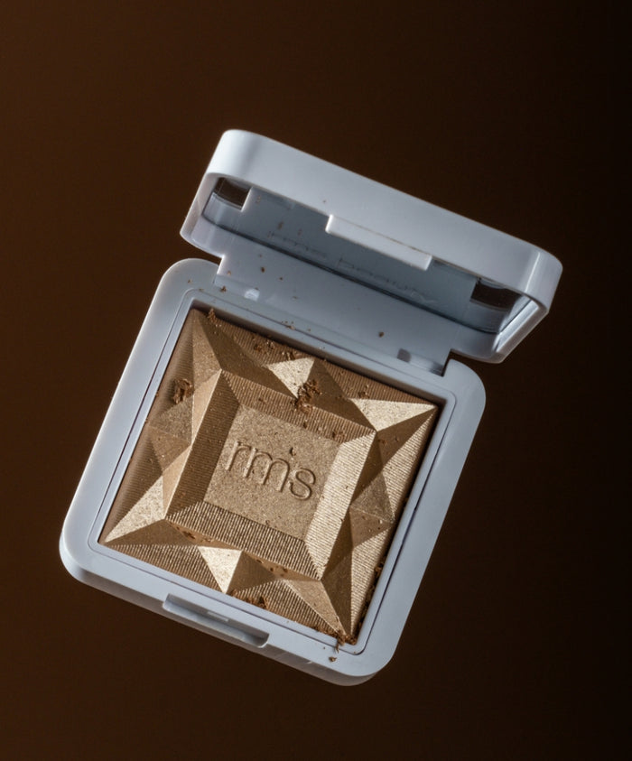 Compact featuring a sculpted golden powder embossed with "rms" in a white case, representing the ReDimension Hydra Dew Luminizer with clean beauty and natural ingredients.