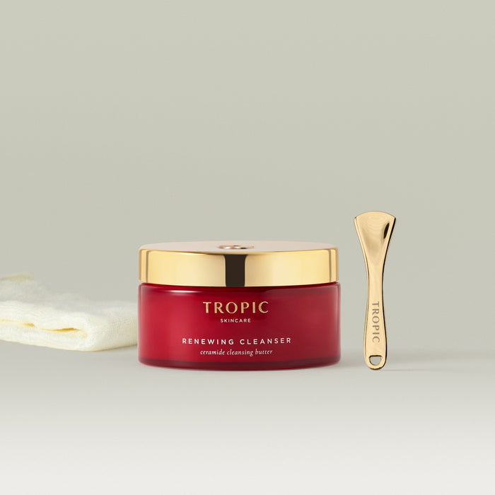 Tropic Renewing Cleanser in a red jar with a gold lid, shown alongside a gold skincare spatula and a soft bamboo cleansing cloth on a neutral background.