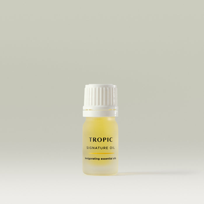 The image shows a small bottle of "Tropic Signature Oil." The bottle is frosted with a golden hue, containing the oil inside. It has a white ribbed cap and a simple, elegant label that mentions "invigorating essential oils." The design is minimalistic and clean.
