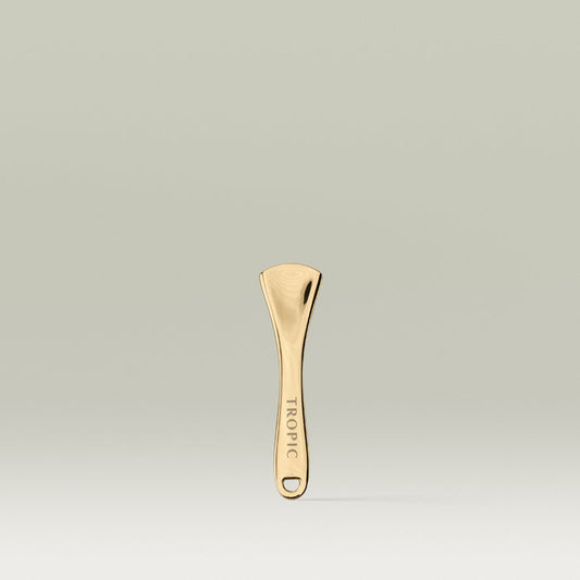 Gold Tropic Skincare spatula standing upright against a soft neutral background, with the word “TROPIC” engraved on the handle.