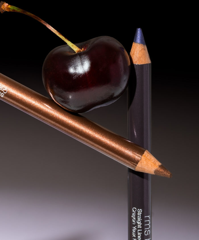 Two sharpened cosmetic pencils, one in dark purple and the other in metallic bronze, balanced next to a ripe dark cherry, highlighting the Straight Line Kohl Eye Pencil in a clean beauty style.