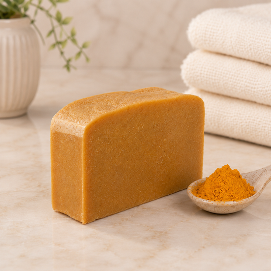 Bar of brown goat milk soap with a spoonful of turmeric on a marble surface with towels and a plant in the background.