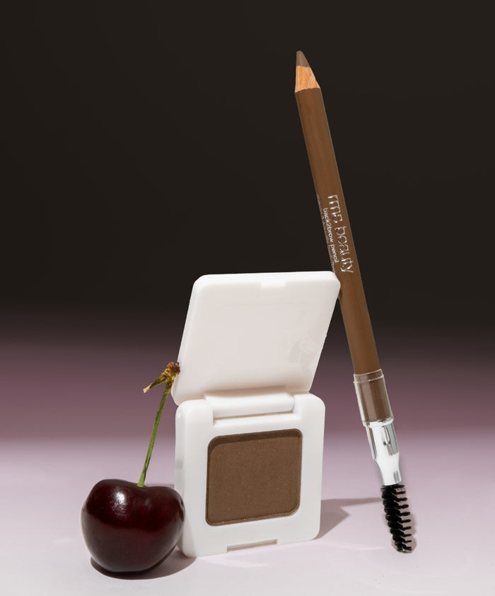 Ultimate Brow Kit featuring a brown eyebrow pencil with spoolie brush and matching powder compact beside a dark cherry, showcasing clean beauty and natural ingredients.
