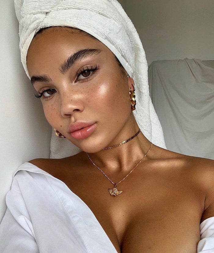 Warm Gold ColourPop SOL Glow Oil illuminates a model’s cheeks, collarbones, and shoulders with a dewy, sun-kissed sheen; towel-wrapped hair, white off-shoulder shirt, and gold jewelry in soft natural light emphasize the glow.