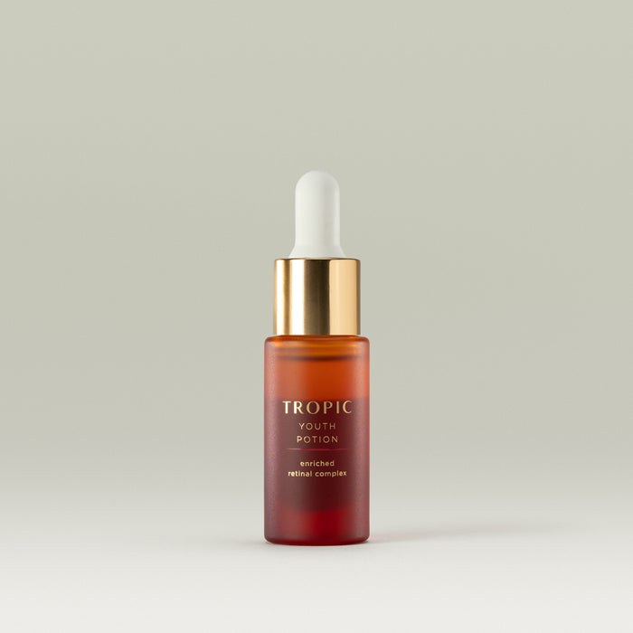 A small glass dropper bottle of Tropic Youth Potion with a gold cap and white pipette, filled with an amber-red serum, standing against a soft neutral background.