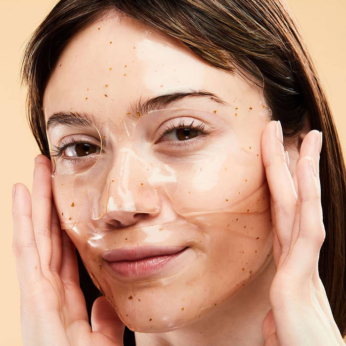 Woman smoothing a clear gold-flecked hydrogel face mask against a warm beige background.