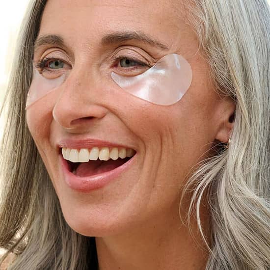 Smiling mature woman with grey hair wearing clear Patchology eye gels for a refreshed and youthful glow.